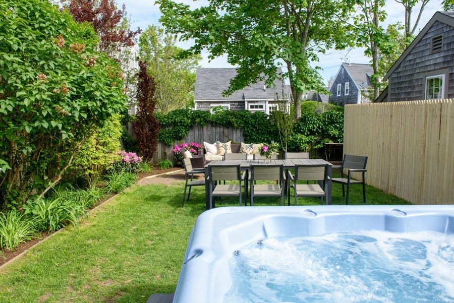 A private backyard features a spacious hot tub in the foreground. Just beyond, a dining table with seating for six is surrounded by greenery and blooming flowers. The scene is framed by a wooden fence and tall trees, creating a serene outdoor setting.