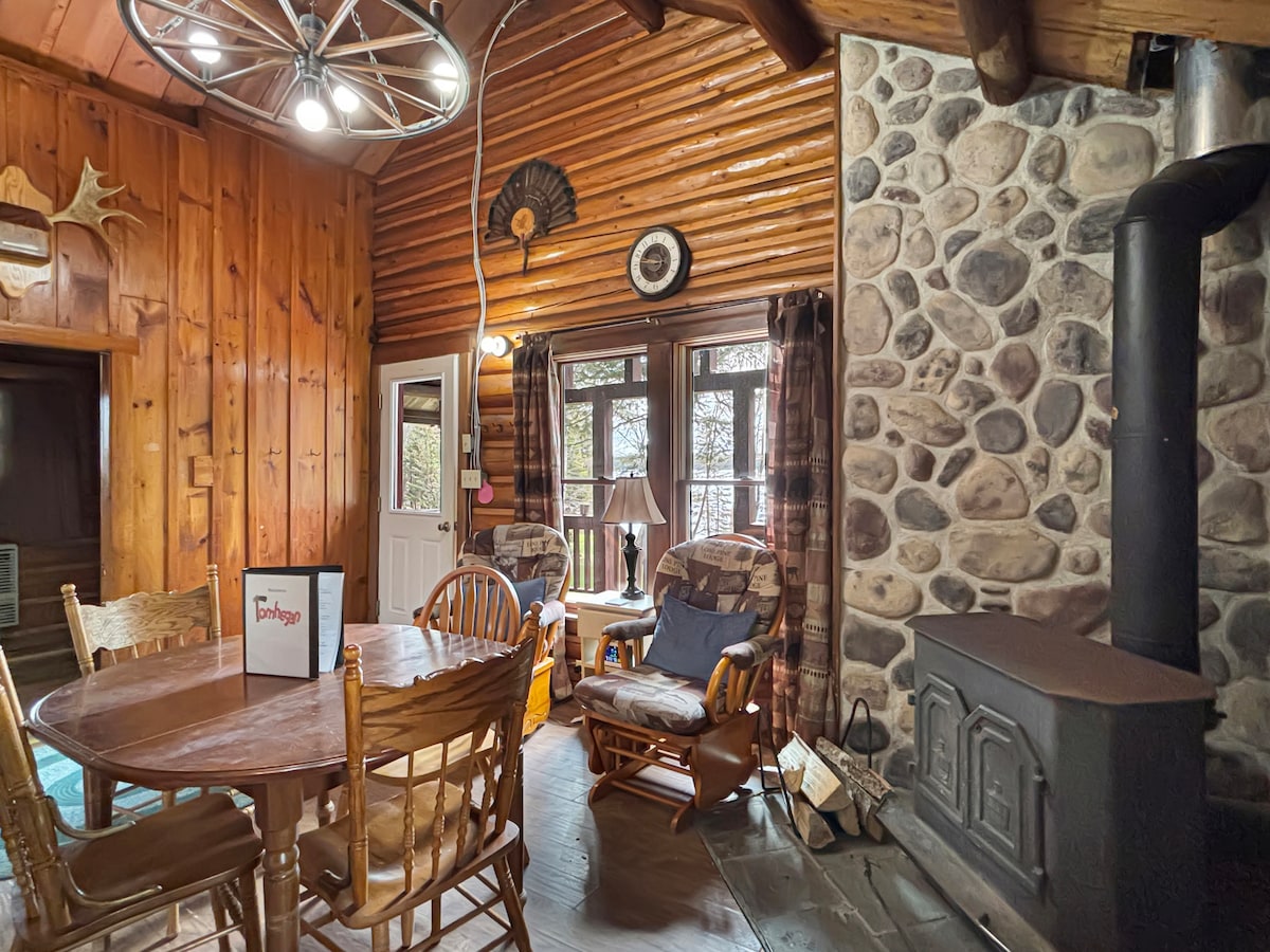 A rustic living space showcases natural wooden walls and a stone fireplace. A round dining table with wooden chairs is situated nearby. Two comfortable chairs are positioned next to the fireplace, inviting relaxation. Large windows allow natural light to fill the space.