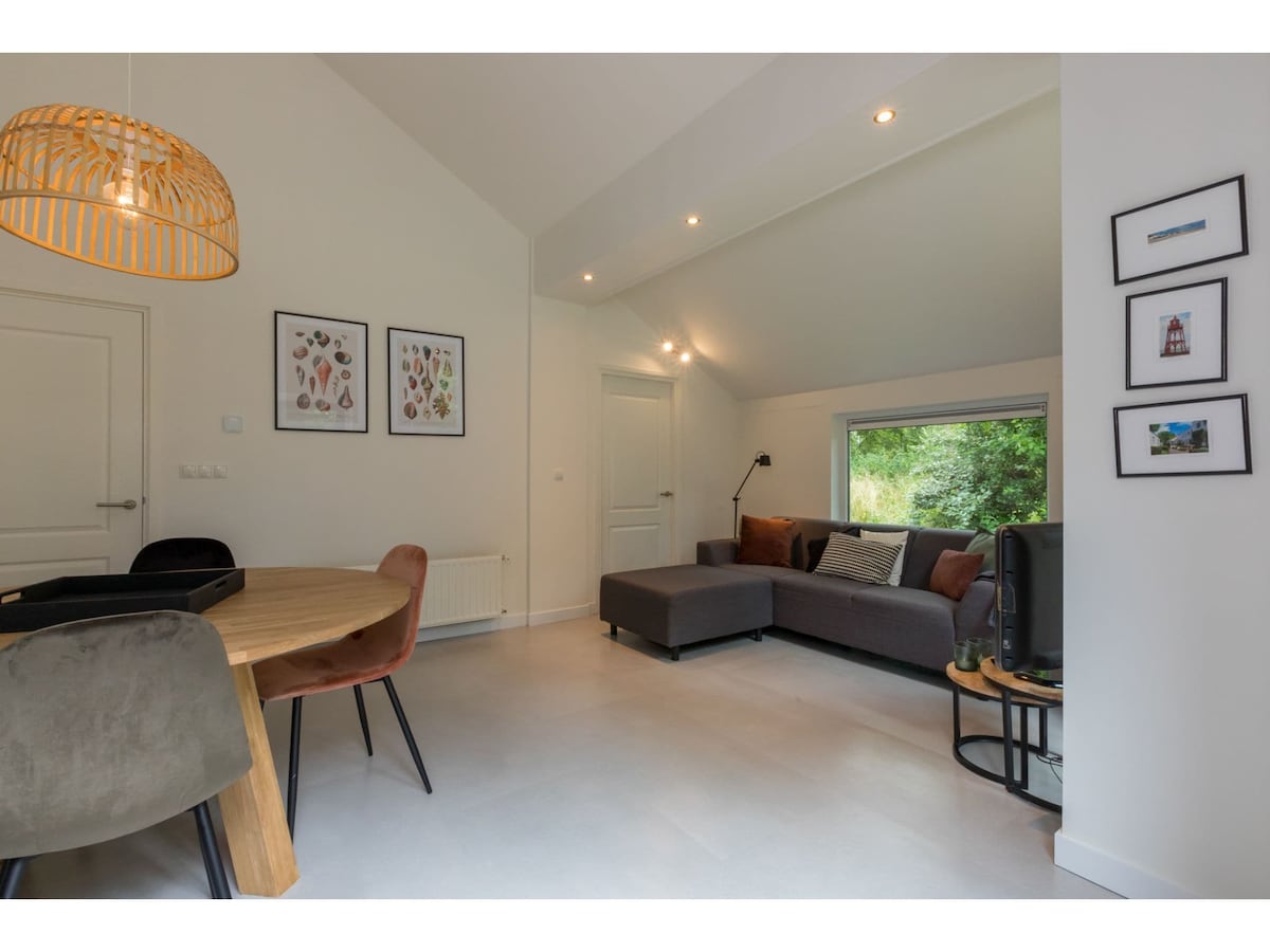 A bright living area is seen, featuring a modern corner sofa and a smart TV. Natural light filters in through a large window, while artwork adorns the walls. A round dining table with stylish chairs connects the living and dining spaces.