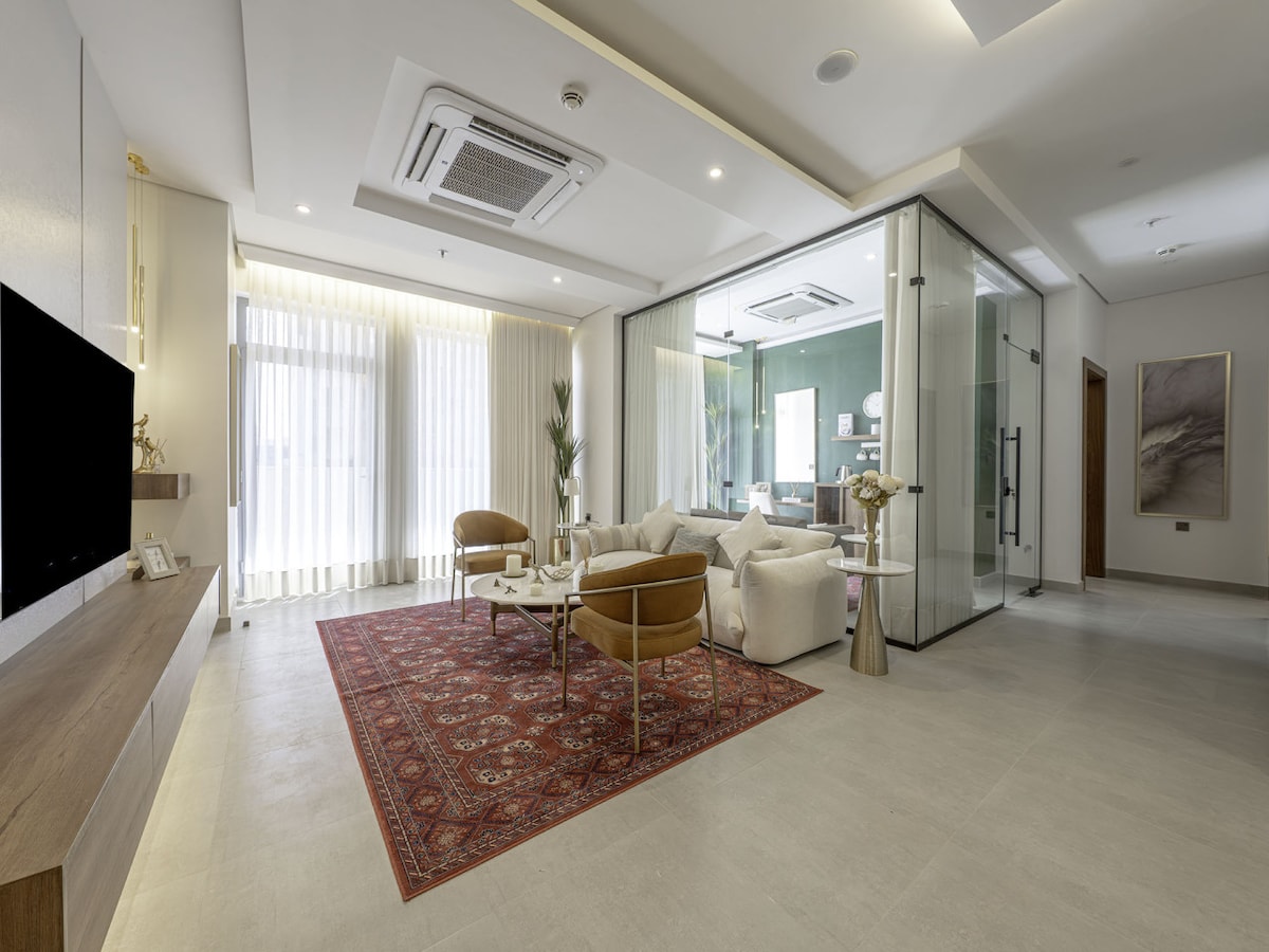 A modern living area features a neutral color palette, highlighted by a patterned area rug. Comfortable seating is provided by two armchairs and a plush sofa. A glass-walled office area is visible, allowing natural light to enhance the space, complemented by sheer window treatments.