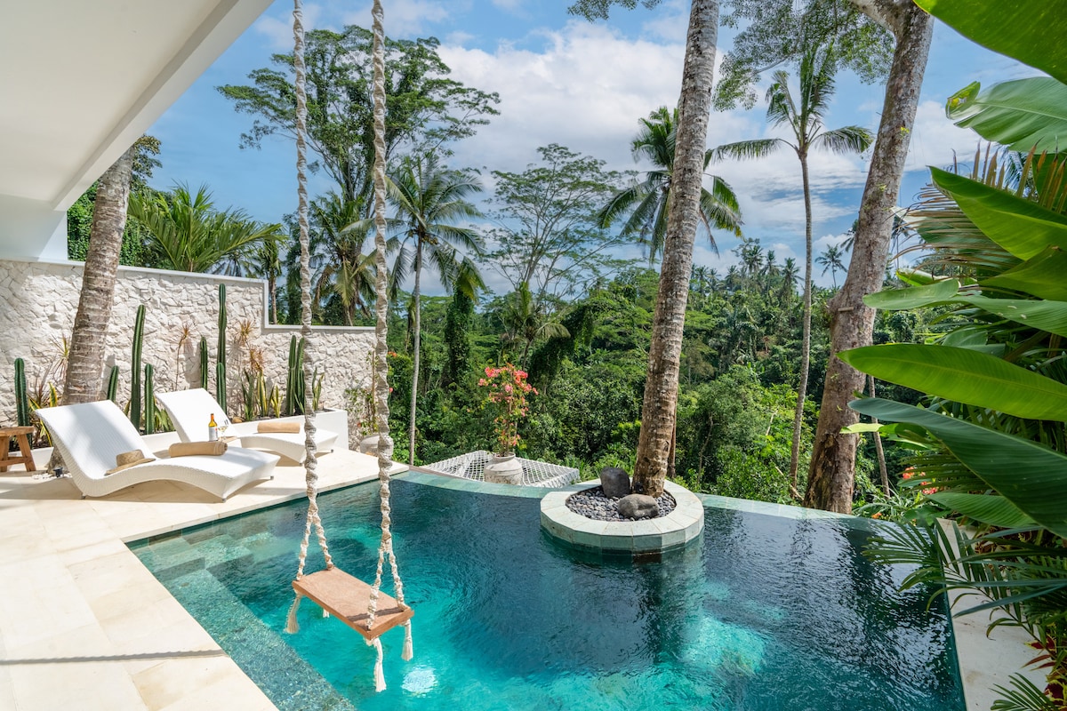 A private swimming pool featuring a natural stone surround is seen in the foreground, with a wooden swing hanging nearby. Two lounge chairs are positioned on the sundeck, offering views of the lush jungle and palm trees that frame the serene landscape.