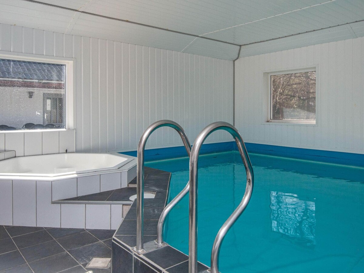An indoor pool area features a spacious swimming pool with a shallow entry step and stainless steel handrails. Adjacent to the pool, a six-person whirlpool is positioned on a tiled platform, while large windows provide natural light to the room.