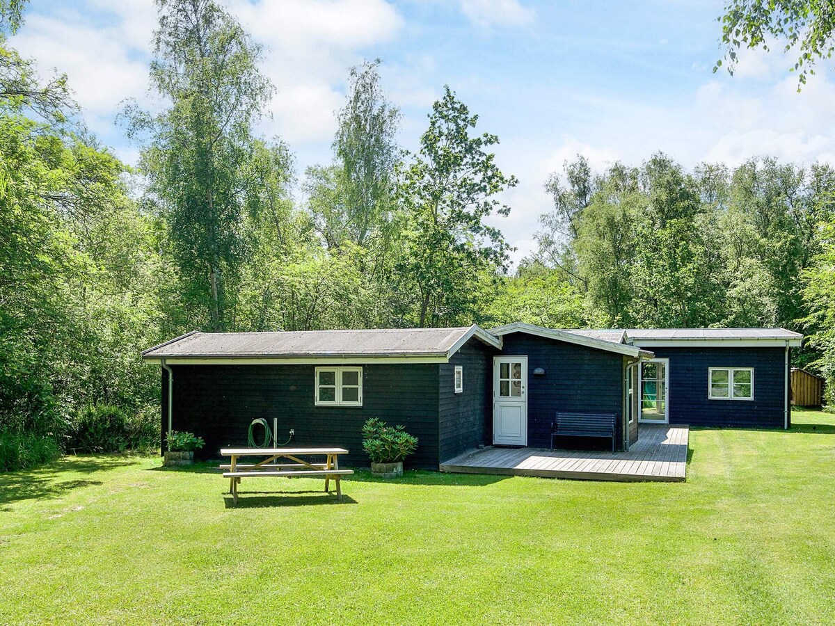 A spacious holiday home is surrounded by lush greenery, featuring a mix of wooden and glass elements. A wooden picnic table is visible on the lawn, providing an outdoor seating area, while the adjacent deck extends from the side of the house.