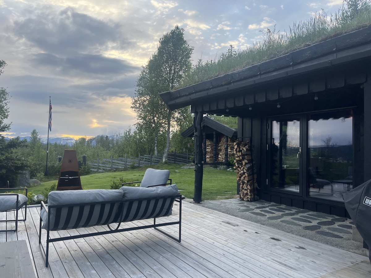 An outdoor seating area is visible on a wooden deck, featuring a gray sofa with plush cushions. A grassy roof tops the cabin, blending harmoniously with the surrounding greenery. Firewood is stacked neatly beside the entrance, and a peaceful landscape is seen under a soft sunset.