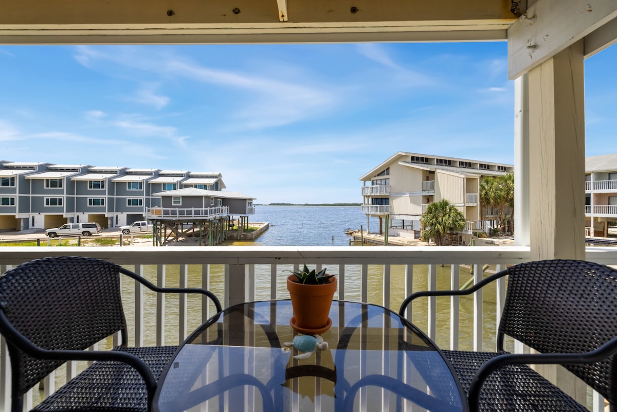 The wrap-around balcony offers a view of calm waters and surrounding buildings. A glass table with two black wicker chairs is positioned prominently, complemented by a small potted plant in the center. The scene is framed by light-colored railing and a clear blue sky.