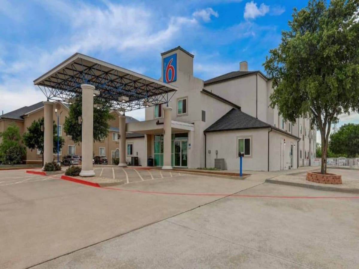 The exterior of the Motel 6 in Weatherford is shown, featuring a covered entrance supported by two columns. A clean parking area is visible, along with landscaping that includes trees and shrubs. The building's facade displays neutral colors, with windows reflecting the sky.