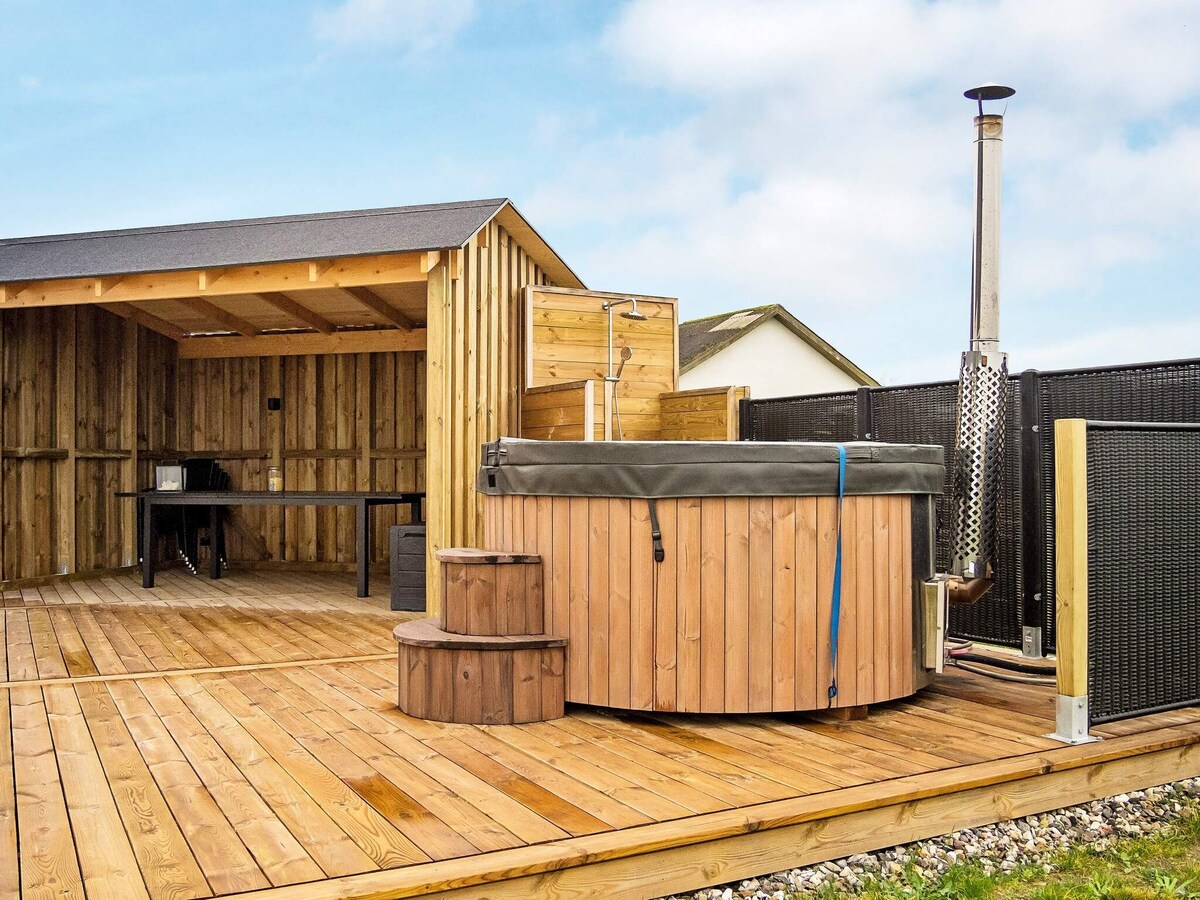 An outdoor wooden deck features a hot tub surrounded by wooden fencing. Adjacent to the tub, a shaded space includes a simple table and chairs. The area is complemented by a rustic structure that provides shelter and additional seating options.