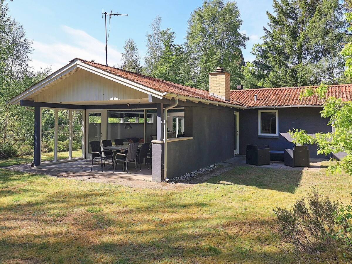 The exterior of the holiday home showcases a single-story structure with a tiled roof and large windows. A covered terrace features an outdoor dining set, surrounded by a green lawn and trees, providing a peaceful view of the natural surroundings.
