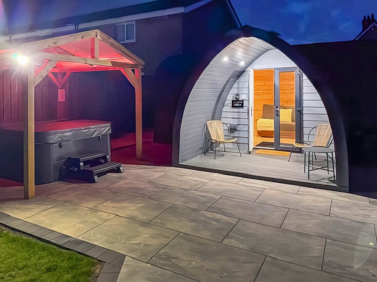 An exterior view of the glamping pod at dusk, featuring a hot tub under a wooden gazebo. Two chairs are positioned outside the pod's entrance, where warm light spills from the interior, highlighting the welcoming entrance.