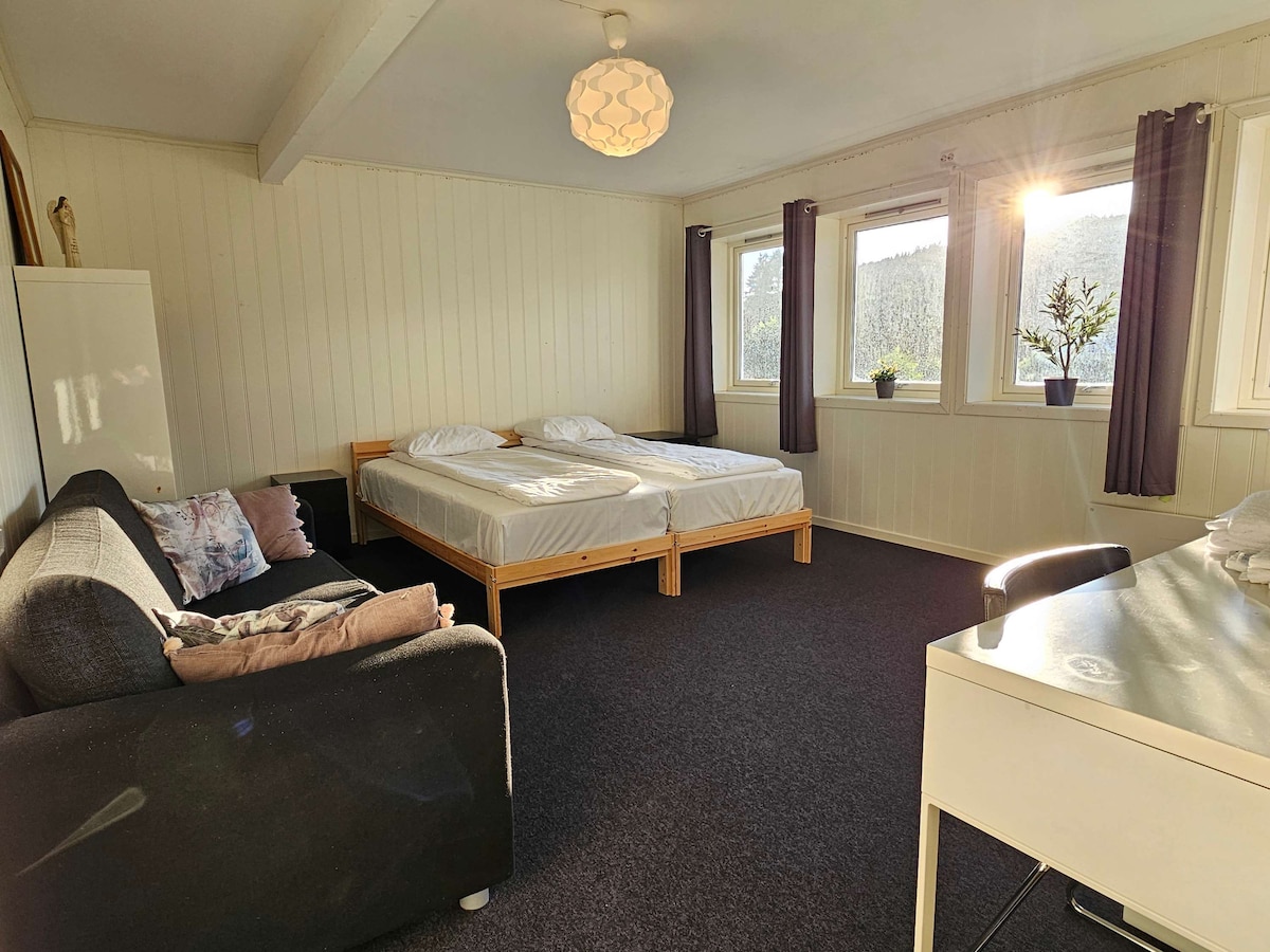 A private room features two spacious beds positioned against the wall, complemented by large windows allowing natural light. A sofa sits in the corner, and a desk is positioned nearby. Light walls and dark carpeting create a relaxed atmosphere.