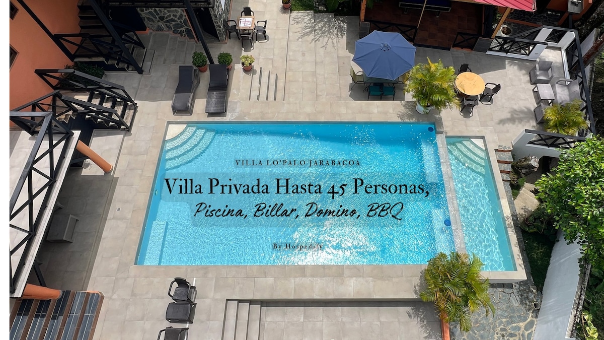 An aerial view of Villa Lo' Palo reveals a spacious outdoor pool surrounded by a tiled patio. Chaise lounges are positioned for sunbathing, and a shaded area with a blue umbrella offers seating. Lush greenery enhances the inviting atmosphere of the outdoor space.