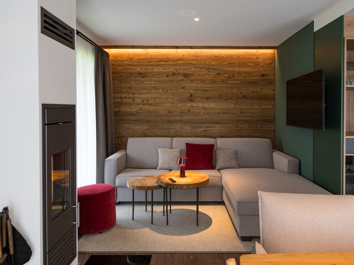 A cozy living area features a grey sofa with a chaise lounge, complemented by a round wooden coffee table and a small red pouf. A wall of natural wood panels adds warmth, while a flat-screen TV is mounted on the wall beside a fireplace, creating a relaxing setting.