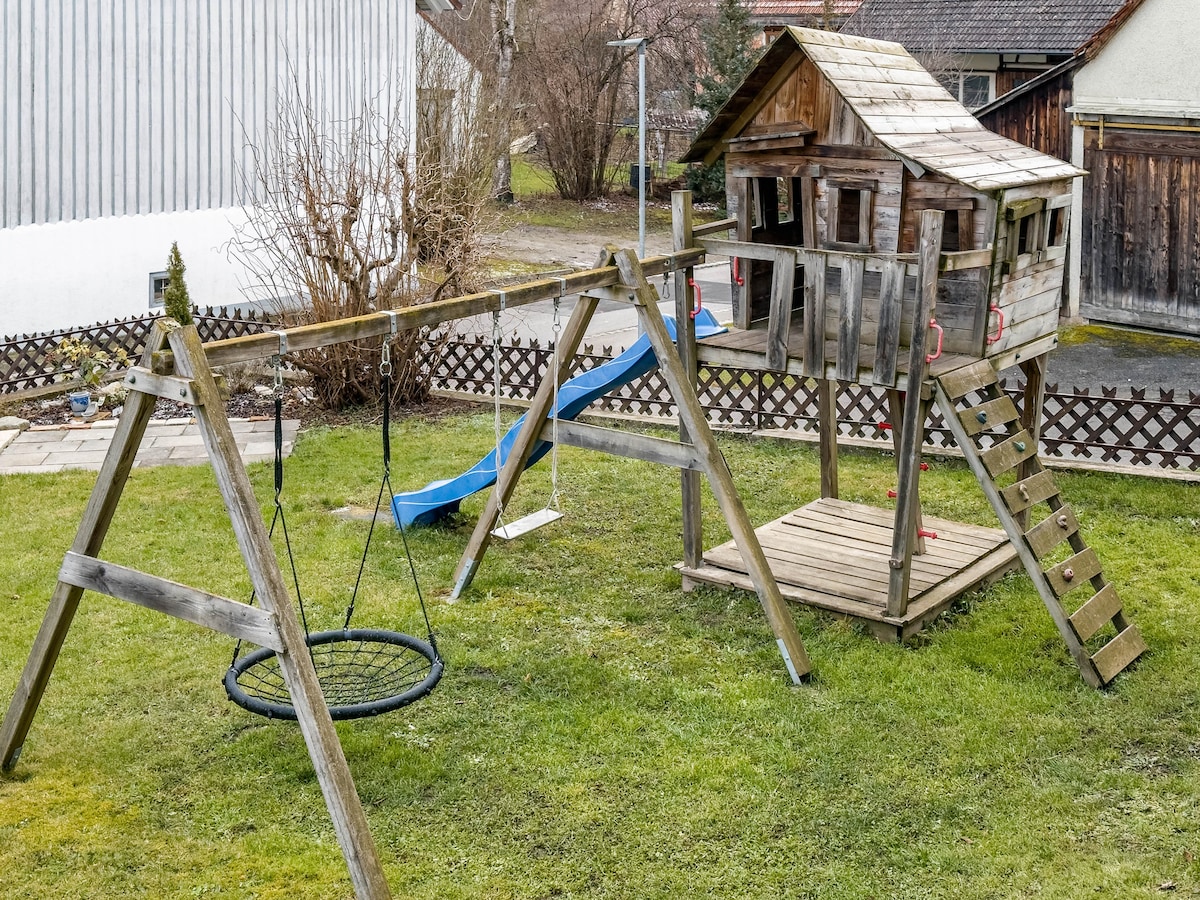 An outdoor play area features a wooden playhouse set atop a platform, accessible by a ladder. Adjacent swings are included, alongside a circular swing. A blue slide extends from the playhouse, with green grass covering the ground surrounding these structures.