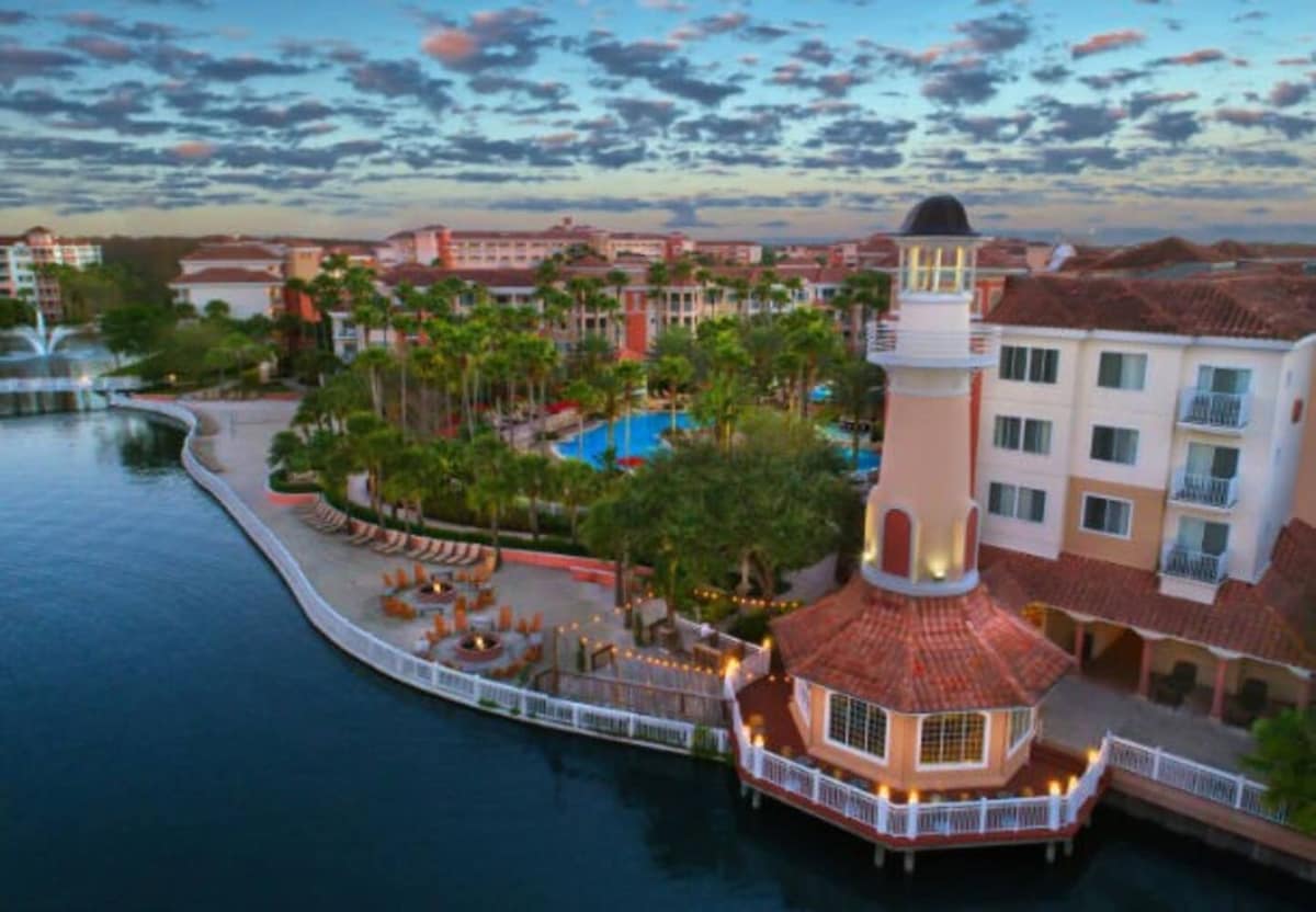 An aerial view showcases the resort's lagoon and lush landscaping. A lighthouse structure stands prominently on the water's edge, while multiple outdoor lounging areas are positioned beside the pool. The sky features scattered clouds and soft colors, creating a serene atmosphere.