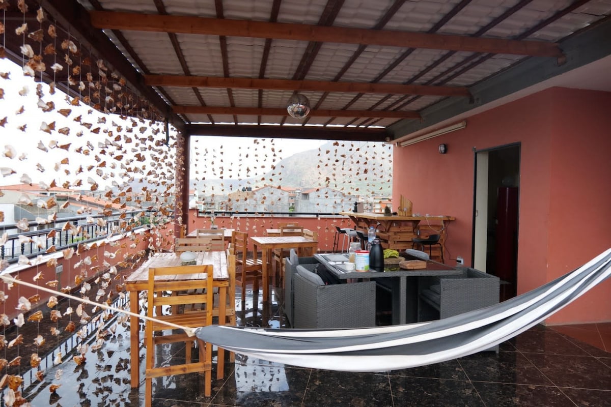 A large rooftop terrace features multiple dining tables and comfortable seating arrangements. A hammock is suspended between two posts, providing a relaxing spot. Decorative elements made of natural materials hang from the overhead structure, adding a unique touch to the open space with a view.