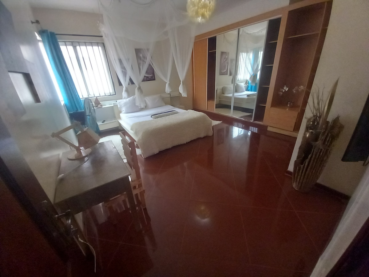 A spacious room features a large bed dressed in soft white linens, draped with a canopy. Natural light filters through the sheer curtains of the window. A mirrored wardrobe and minimalist furniture contribute to a serene environment, complemented by glossy tiled flooring.