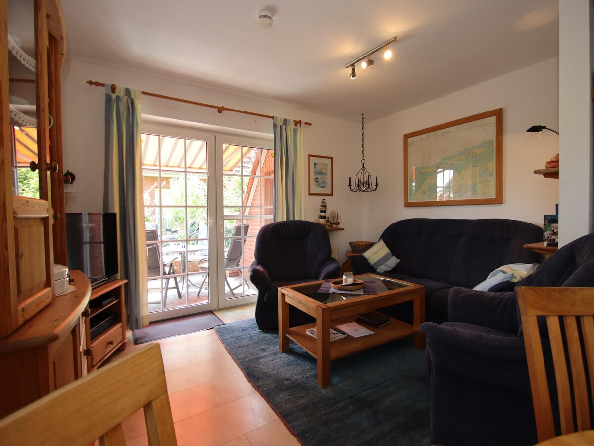 The living area features comfortable seating arrangements with two dark armchairs and a spacious sofa, arranged around a wooden coffee table. Large glass doors lead to the outdoor terrace, allowing natural light to fill the space. A warm rug complements the seating area.