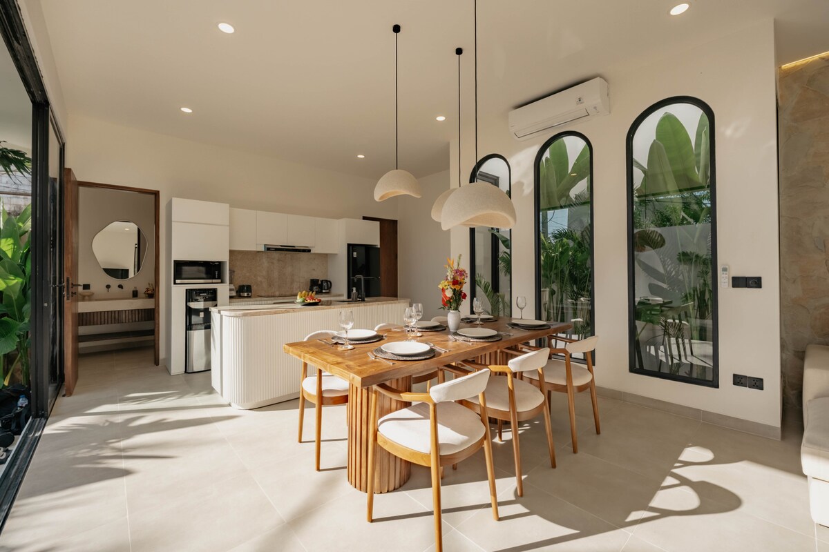 A modern dining area features a wooden table laid with place settings for four. Three pendant lights hang above, providing gentle illumination. Large arched windows frame views of greenery, enhancing the light and open feel of the space.