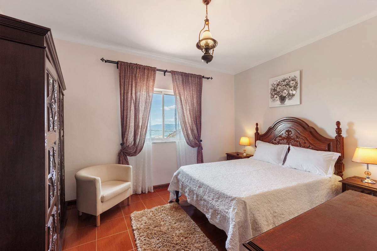 The bedroom features a spacious bed adorned with a light-colored quilt. A wooden wardrobe and nightstands are positioned beside the bed, creating a harmonious setup. Natural light enters through a window, framed by sheer curtains, while a soft rug adds warmth to the tiled flooring.