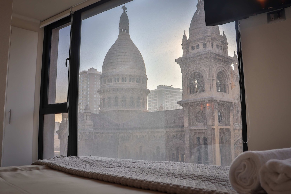 A large window offers views of a historic building with distinctive domes, framed by a cozy sleeping area. The soft tones of the bed linens complement the architectural details visible outside. Light filters through the glass, creating a serene atmosphere.