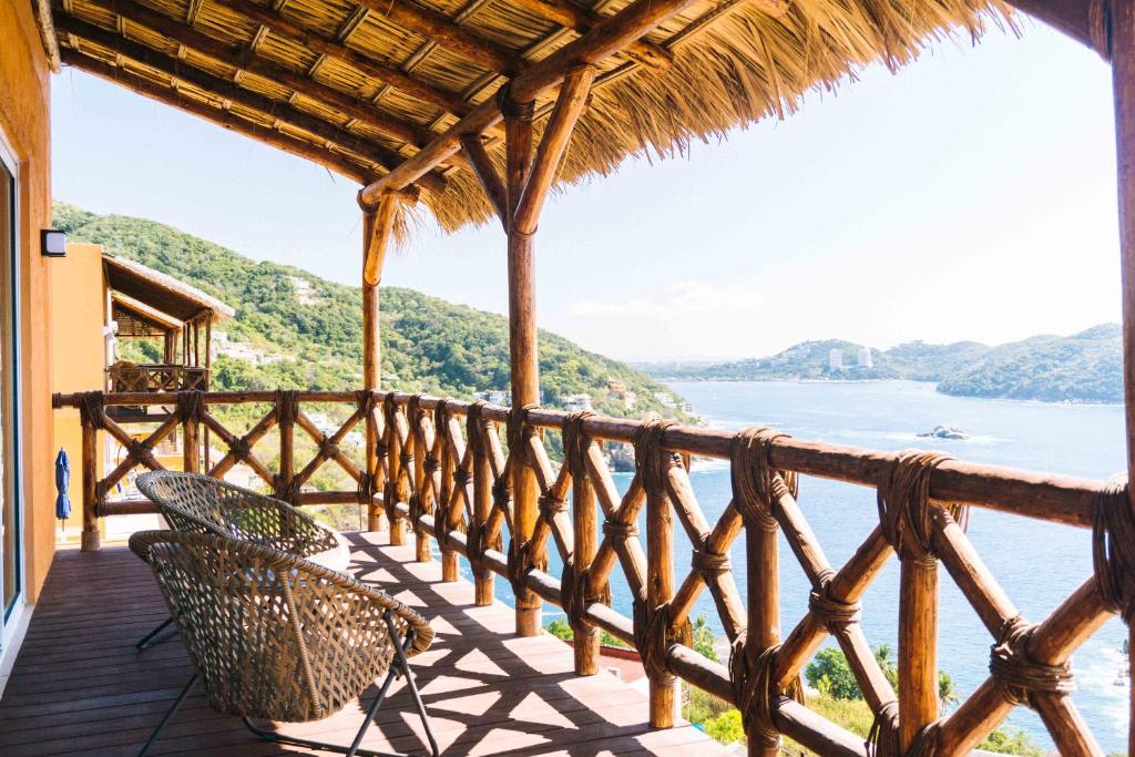 A covered terrace features rustic wooden beams and a thatched roof, offering stunning views of the coastline. Two woven chairs and a table are positioned to enjoy the serene outdoor setting, surrounded by lush green hills and sparkling water.