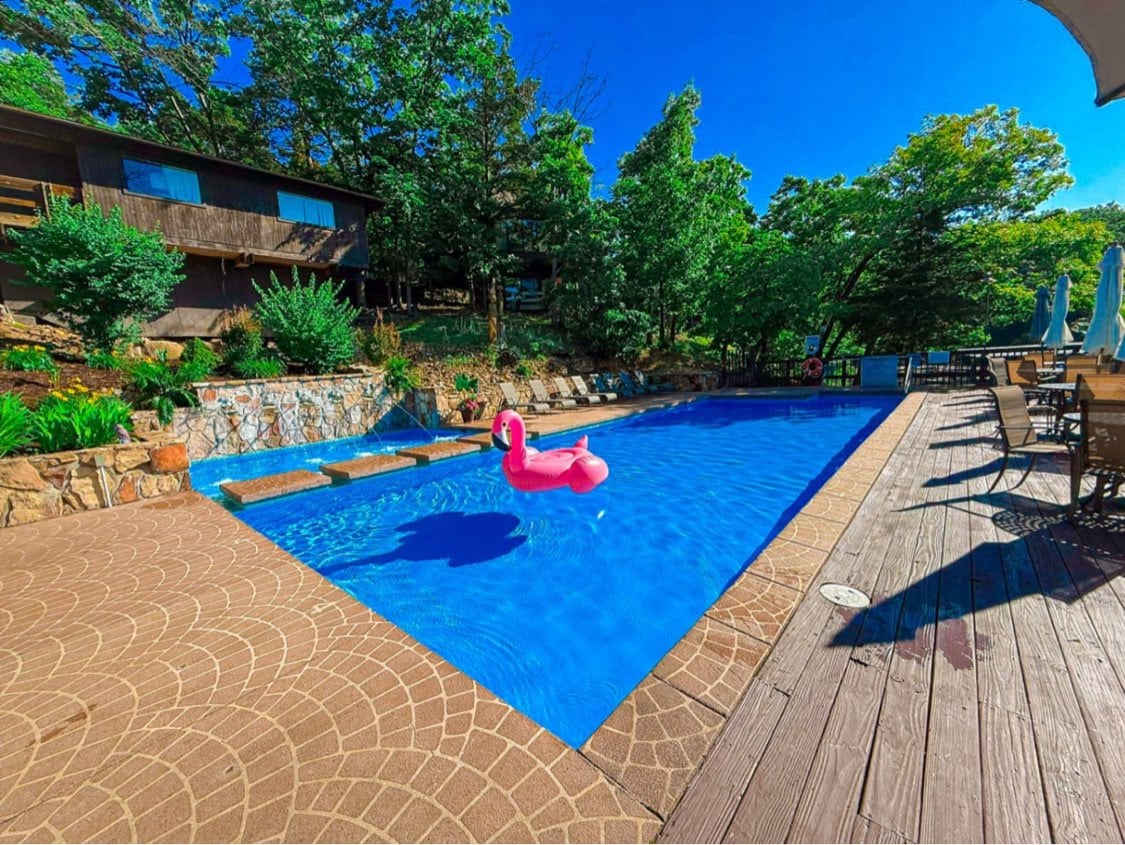 A refreshing outdoor swimming pool is surrounded by lush greenery and wooden decking. A pink inflatable float is positioned in the center of the clear blue water, complementing the inviting space. Comfortable lounge chairs are arranged along one side of the pool, offering relaxation options.
