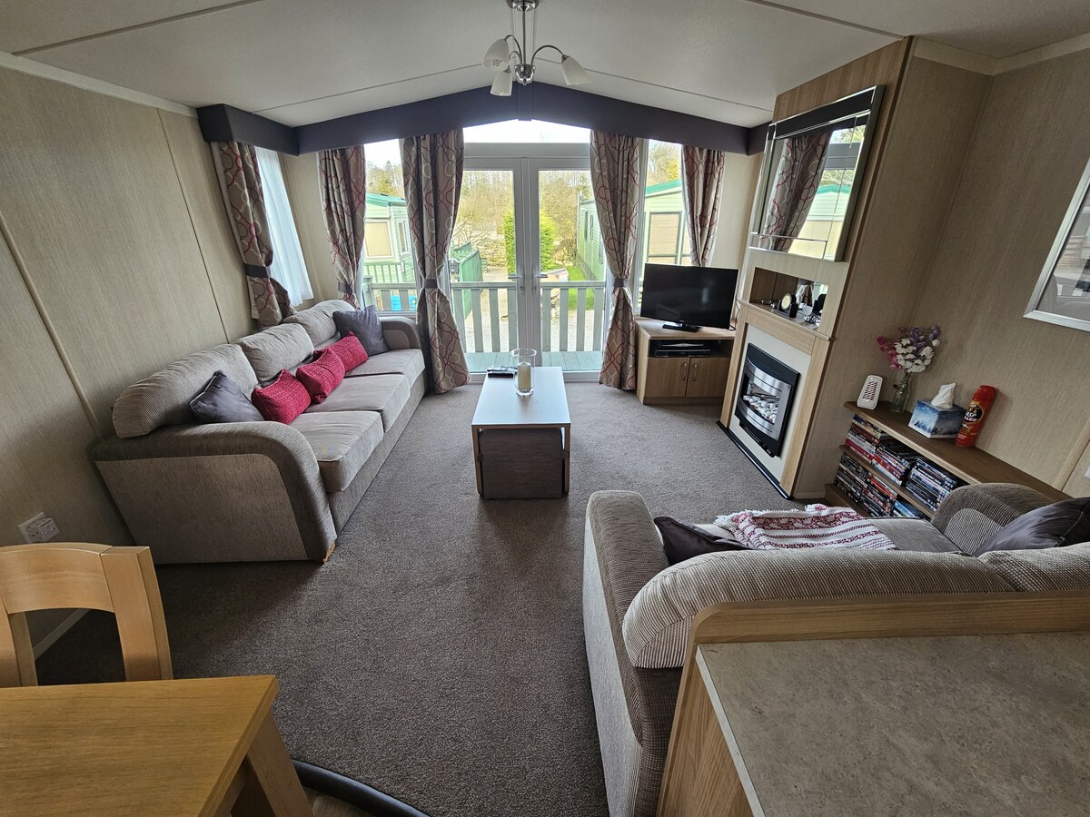A spacious living area includes comfortable seating arranged around a coffee table. Large windows allow natural light to fill the room, with a view of the outdoors. A television is positioned for easy viewing, complemented by a decorative fireplace and shelving with various items.