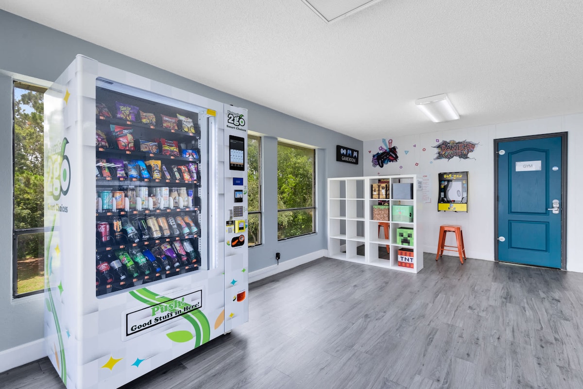 A well-lit room features a vending machine stocked with various snacks and beverages. Shelving units are arranged against one wall, displaying games and additional items. Large windows allow natural light to illuminate the space, complementing the modern decor and colorful art on the walls.