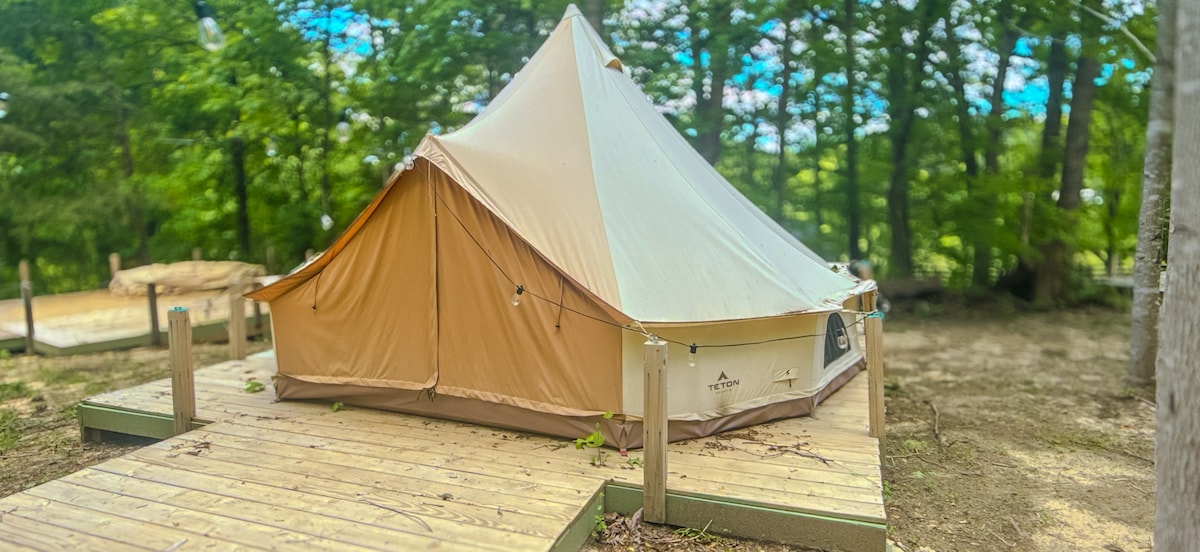 A 16-foot canvas yurt sits on a wooden deck surrounded by lush greenery. The structure features a beige and light brown exterior, blending with the natural setting. A clear sky is visible above, and pathways lead from the yurt to the nearby forest.