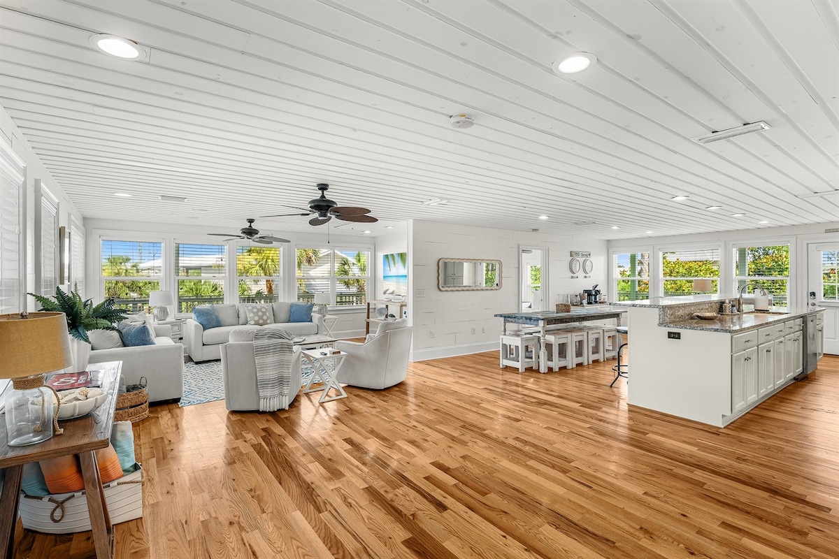 The spacious living area features a light wood floor complemented by a white beamed ceiling. A comfortable seating arrangement is present along with a dining area. Large windows allow natural light and showcase views of greenery and the outdoors.
