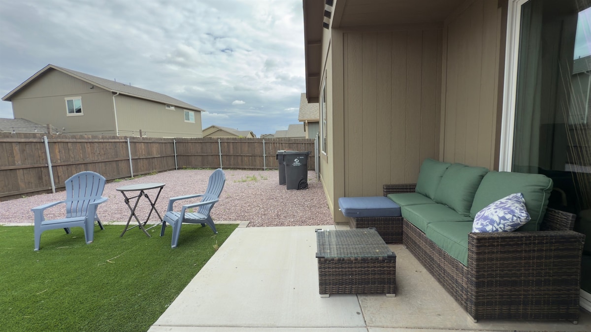 An outdoor seating area features a green cushioned sofa and a wicker table, complemented by two blue Adirondack chairs. A small café table is positioned nearby, and a paved patio area is set against a backdrop of a gravel yard and fenced surroundings.