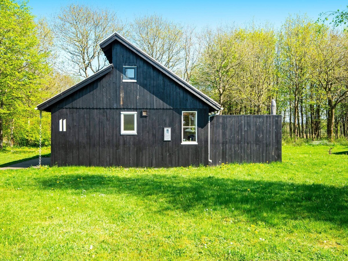 A black wooden holiday home is set against a backdrop of lush green grass and trees. Large windows allow natural light to fill the interior, while a section of the yard features a wooden fence. A swing hangs nearby, enhancing the outdoor space.