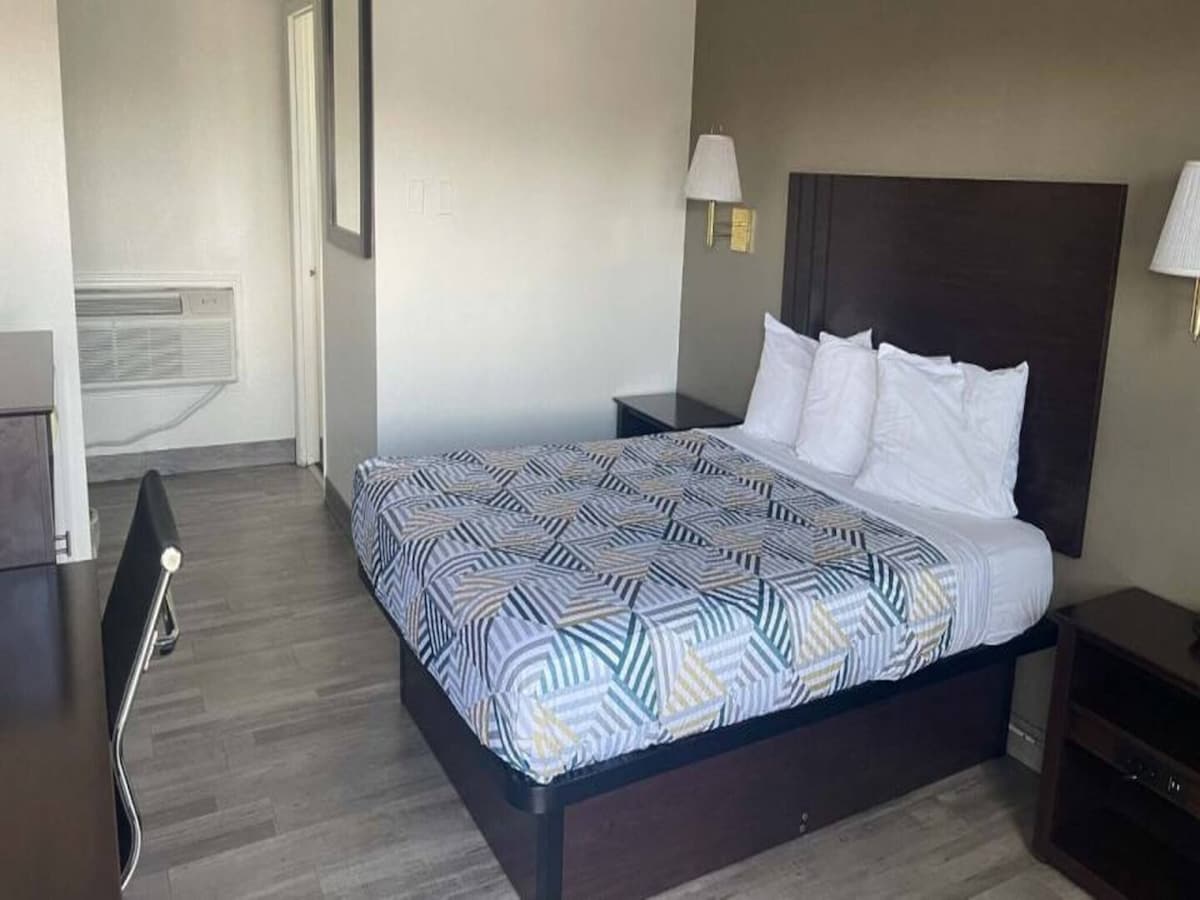 A clean and comfortable room features a spacious bed adorned with patterned bedding. A desk and chair are positioned next to a low table, and soft lighting is provided by wall-mounted lamps. A window allows natural light to brighten the space, with an air conditioning unit visible on the wall.