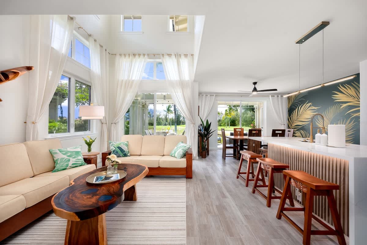 A bright living area showcases large windows with white curtains allowing natural light to fill the space. A seating arrangement includes a light-colored sofa and a wooden coffee table. The dining area features a modern table with high stools, backed by a tropical-themed wall.