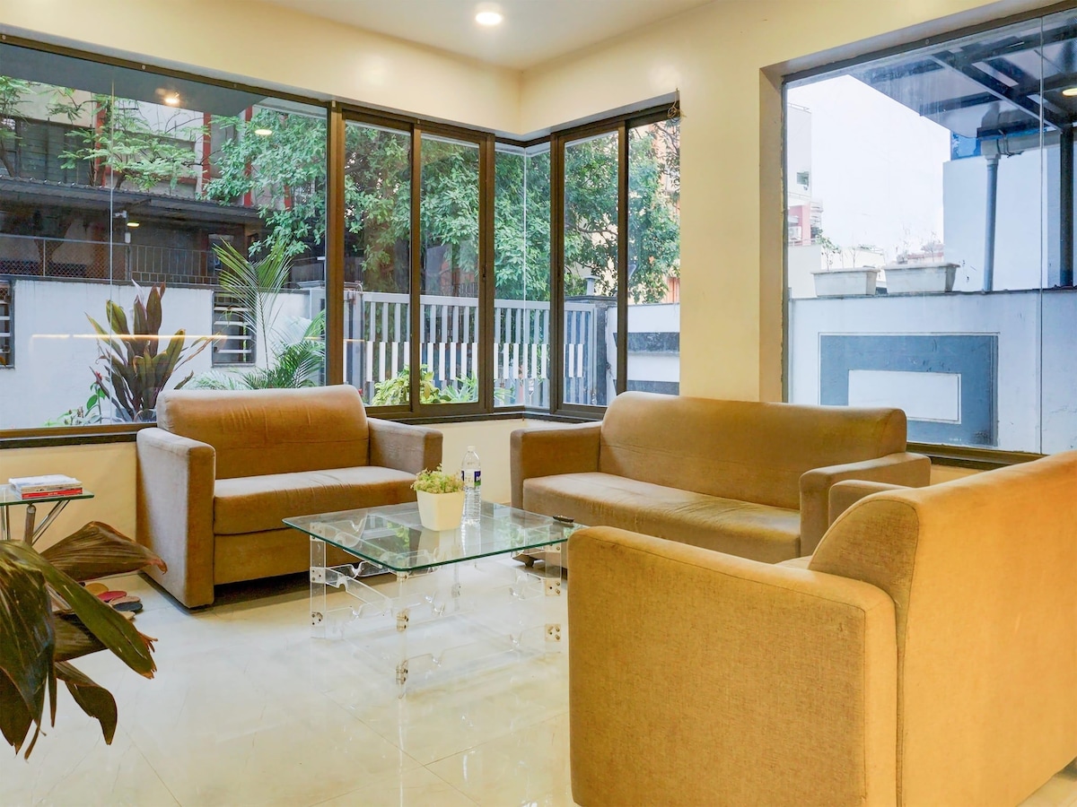 A bright communal lounge area features large windows that offer abundant natural light. Two comfortable sofas and a glass coffee table are arranged on a tiled floor, complemented by indoor plants adding a touch of greenery. A welcoming atmosphere is presented for relaxation and socializing.