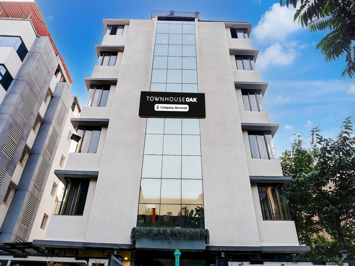 A modern building featuring a geometric facade with large glass windows allowing natural light to fill the interior. The establishment is adorned with greenery at its base, enhancing the contemporary design. A clear blue sky forms the backdrop, adding to the setting.