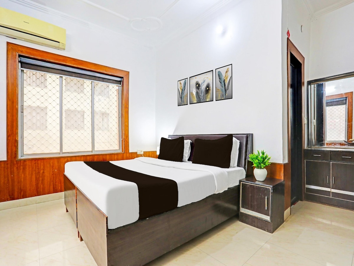 A comfortably appointed room features a spacious bed with dark accents and light bedding. Artwork depicting floral themes is displayed above the bed. Natural light filters in through a large window, with a complementary side table and a dresser enhancing the room's functionality.
