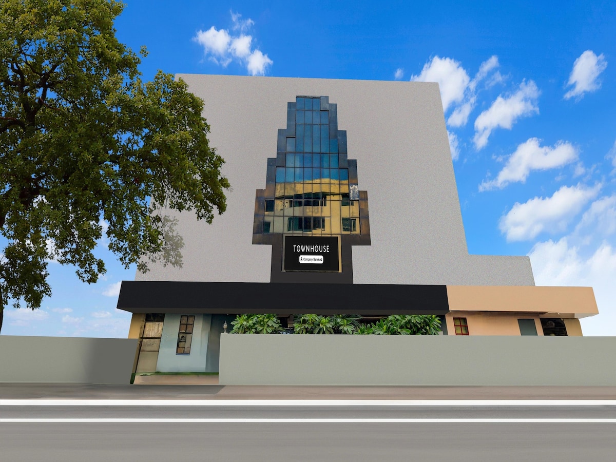The exterior of the building features a modern design with a prominent geometric facade. Large glass windows reflect the sky, contributing to a spacious appearance. A welcoming entrance is highlighted by greenery and a clear sign indicating the location, set against a bright blue sky.