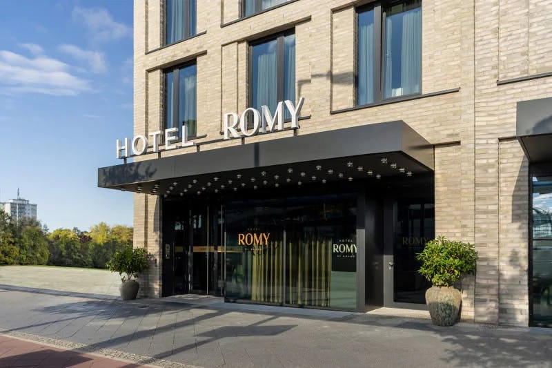 The hotel entrance features a modern facade with a combination of large glass windows and textured brick walls. Above the entrance, the name 'Hotel ROMY' is displayed prominently. Flanking the entrance are large, decorative planters with lush greenery.