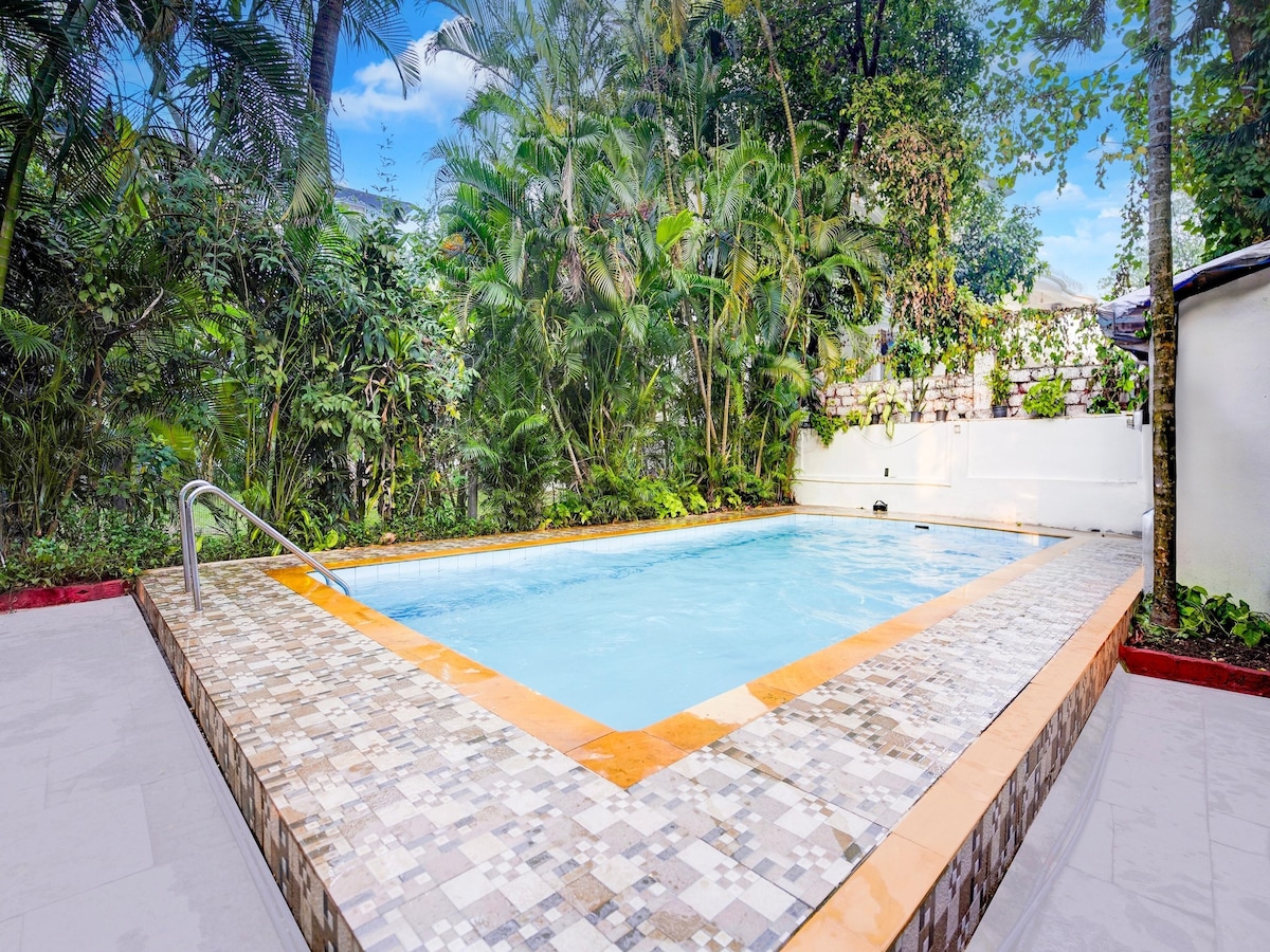 A seasonal outdoor swimming pool is framed by tropical greenery, with a smooth surface reflecting the sky. The pool's tiled edges incorporate warm orange and neutral tones, enhancing the serene environment for relaxation.