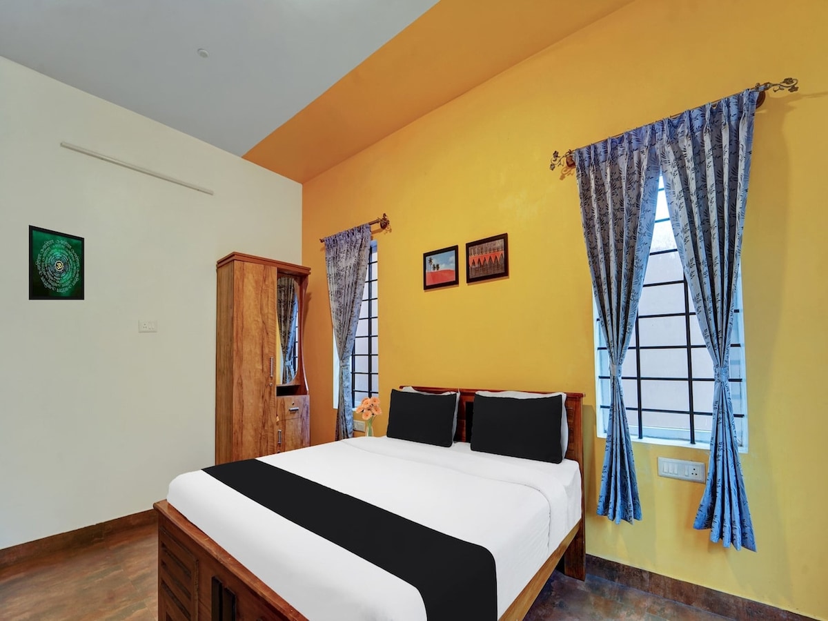 A bright bedroom features a spacious bed with a black and white duvet cover. Wooden furniture includes a wardrobe, and artwork adorns the walls. Natural light enters through two windows, draped with patterned blue curtains, complementing the yellow walls.