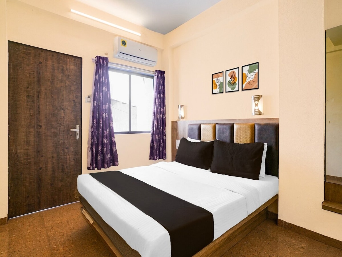 A spacious bedroom is furnished with a large bed featuring a black and white bedding set. The room includes a window dressed with purple curtains and decorative wall art. A door and an air-conditioning unit are visible, contributing to a comfortable atmosphere.
