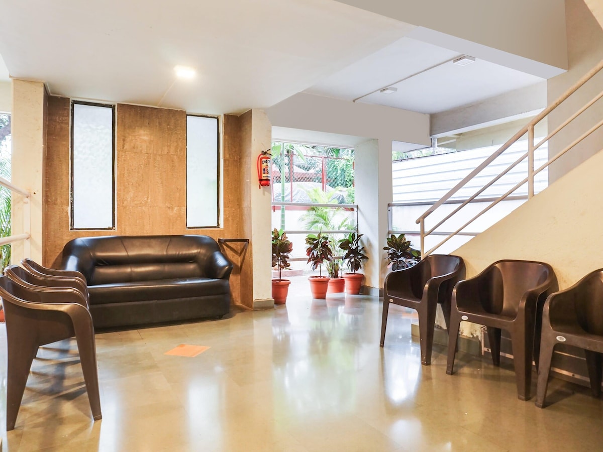 A shared lounge area features a black sofa complemented by several brown chairs. Potted plants are situated near large windows, allowing natural light to enhance the space. A staircase with a simple railing is visible in the background, providing additional entry to the area.