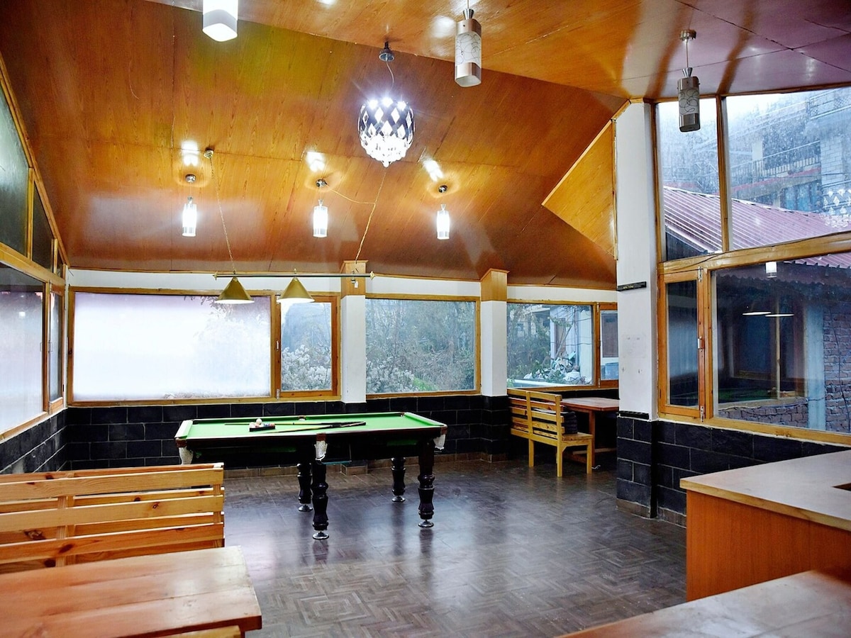 A spacious common area features large windows allowing natural light to illuminate the room. A pool table occupies the center, surrounded by wooden seating. The ceiling is adorned with pendant lights, enhancing the welcoming ambiance of the space.
