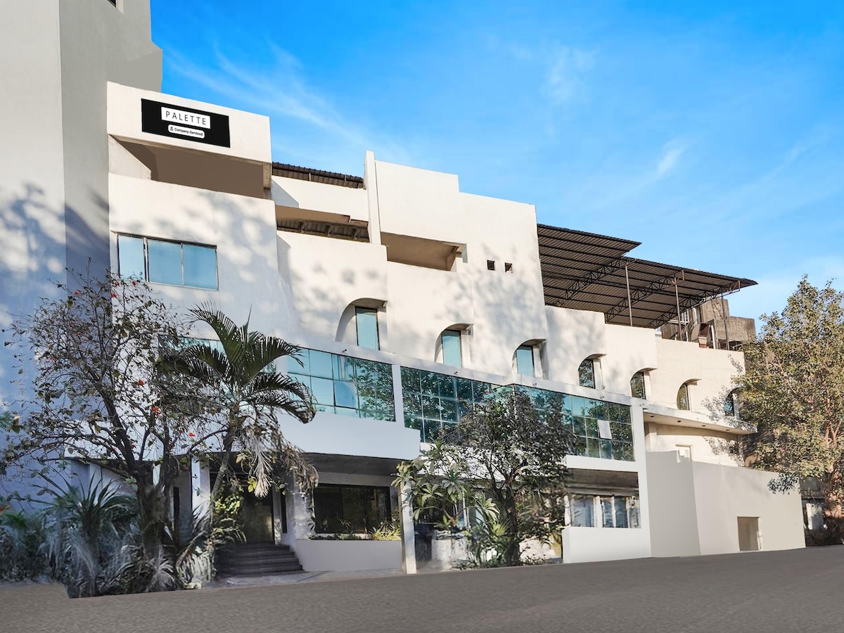 The exterior of the building features a modern design with multiple levels and large windows. Lush greenery surrounds the entrance, enhancing the welcoming atmosphere. A clear blue sky provides a bright backdrop, highlighting the structure's clean lines and architectural features.