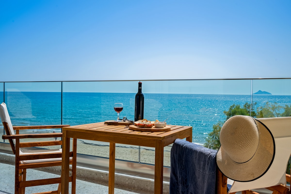 A wooden table is set for two on a private terrace, overlooking the sparkling blue sea. A bottle of wine and two glasses are placed alongside a plate of fresh food. Comfortable wooden chairs provide a serene spot for enjoying meals with a coastal view.