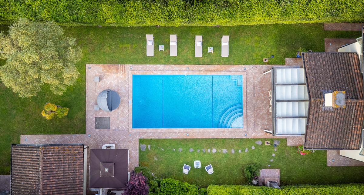 An aerial view of a private pool surrounded by a grassy area, showcasing several sun loungers positioned neatly along the pool's edge. The pool appears clear and inviting, with a gentle ripple on its surface. Surrounding trees and shrubs create a sense of privacy.