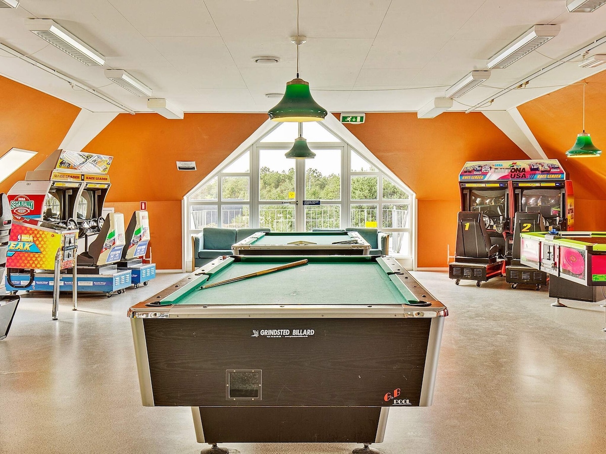A vibrant recreational space features a green billiard table at the center. Bright orange walls are complemented by large windows that provide natural light. Various arcade games are positioned along the walls, offering entertainment options for guests.