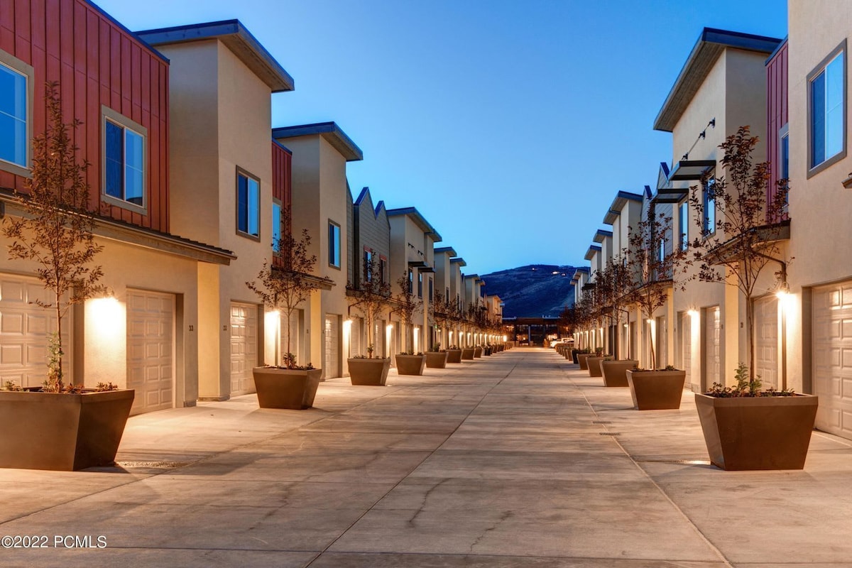 A neatly arranged row of townhouses is illuminated by warm lighting along a wide, paved pathway. Large planters with trees decorate the sidewalks, enhancing the community feel. In the background, distant hills are visible under a dusky blue sky.