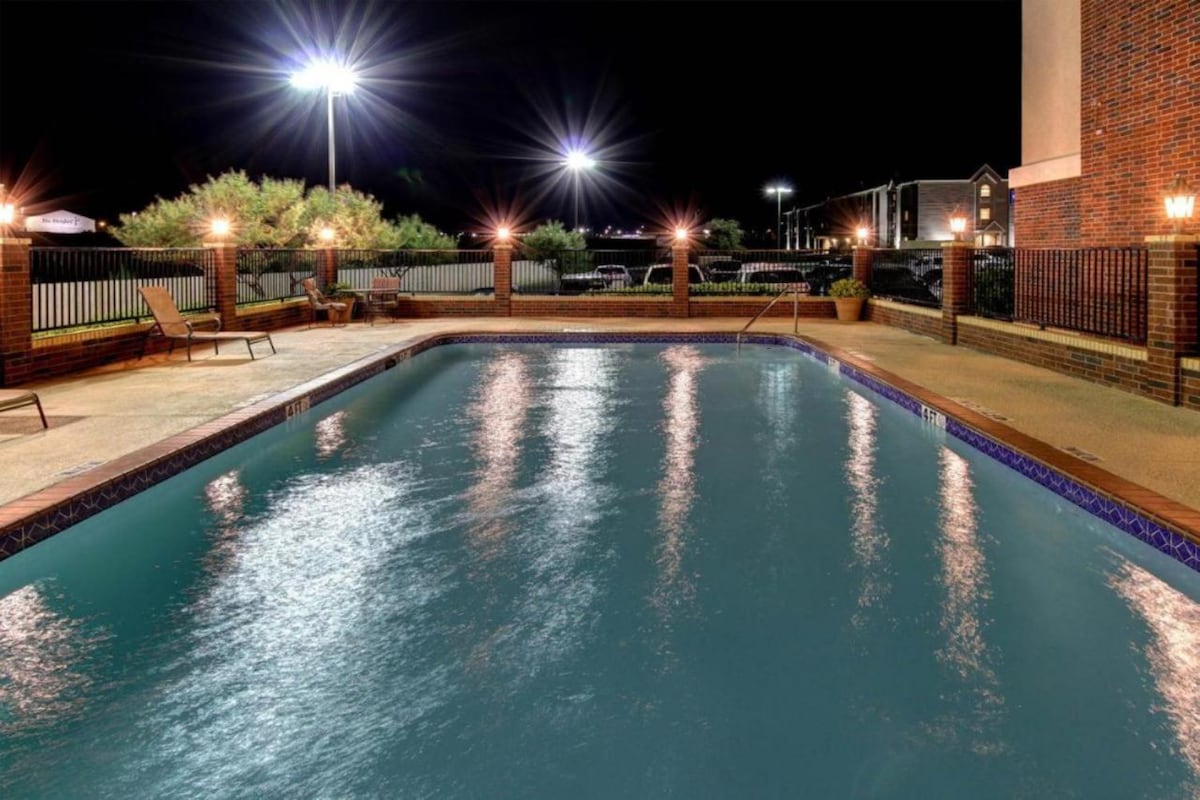 An outdoor swimming pool is illuminated at night, featuring bright lights surrounding the area. Lounge chairs are positioned nearby on the textured patio surface. The water reflects the light, creating a serene atmosphere for evening relaxation.